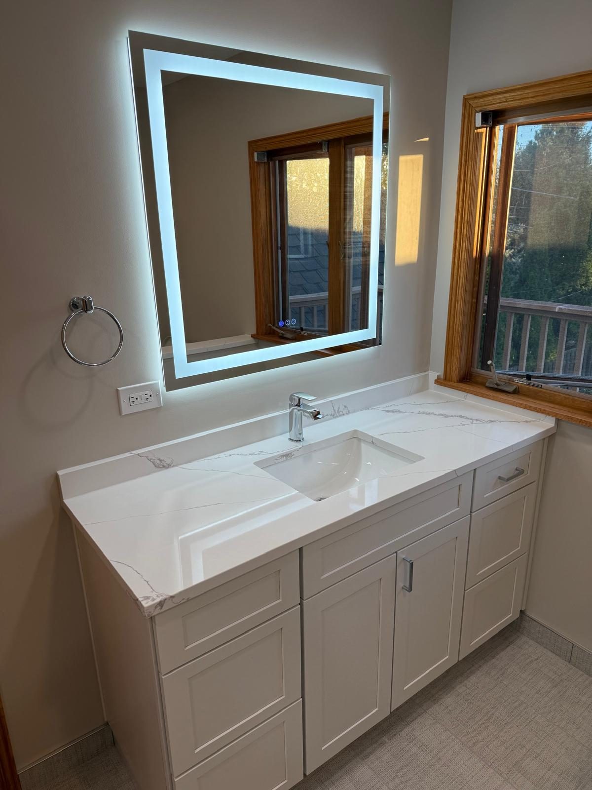 White vanity with stone-look top and a large LED backlit mirror beside a window