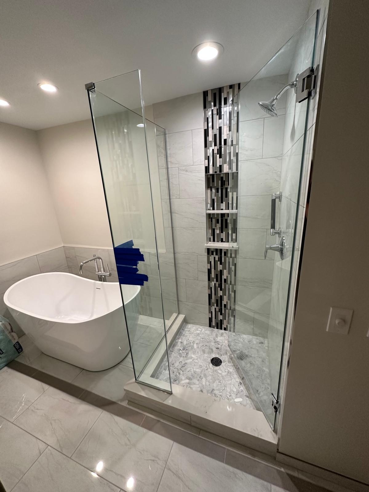 Freestanding white soaking tub next to a frameless glass shower with a mosaic accent strip